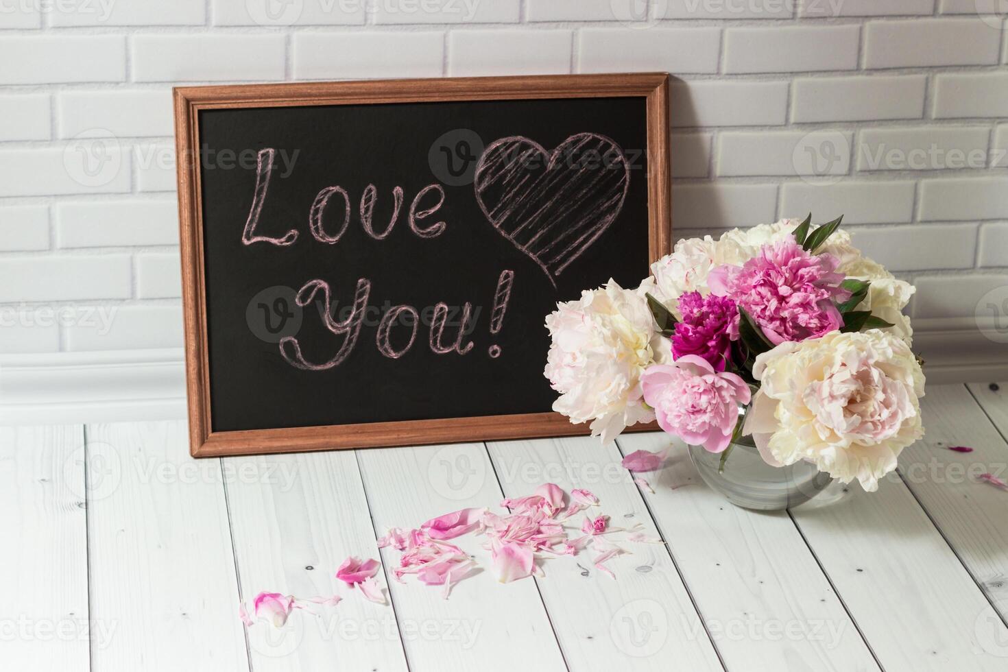 Beautiful bouquet of pink and white peonies in the glass vase with petals and black chalkboard with inscription Love You on the light grey brick and wood background photo