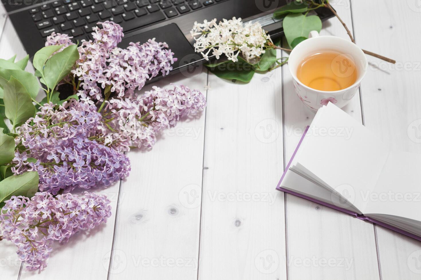 Beautiful branch of white and violet lilac flowers with opened pad and black opened laptop, lying on the white wooden background with cup of tea, mock up perspective view photo
