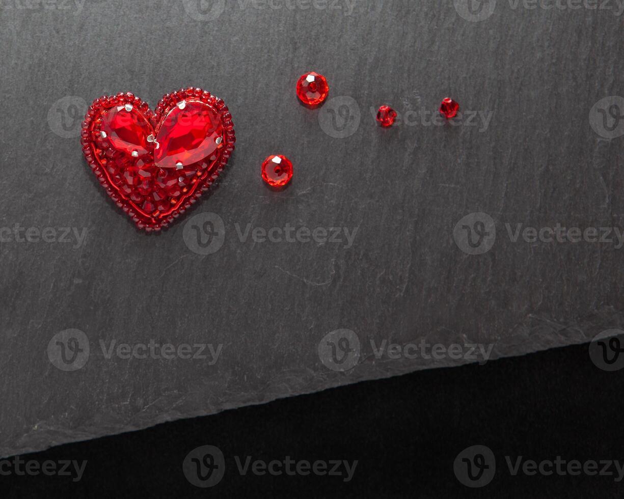 Beautiful handmade embroidered red heart brooch with crystals,lying flat on the dark gray stone rectangular background, horizontal top view with empty place for text photo