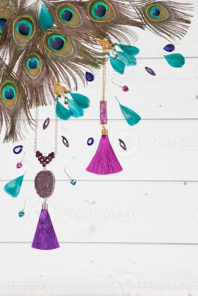 Handmade turquoise and violet bijouterie with gems, tassels and peacock feathers in boho style, lying flat on the white wooden vertical background, top view, diagonal composition. photo