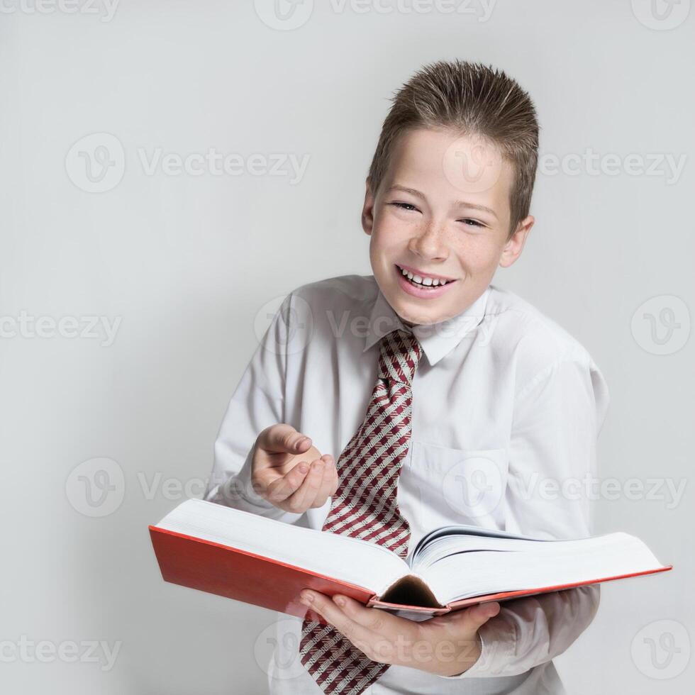 The nice smiling boy teenager in a white shirt and a tie reads a big red funny book on a gray background photo