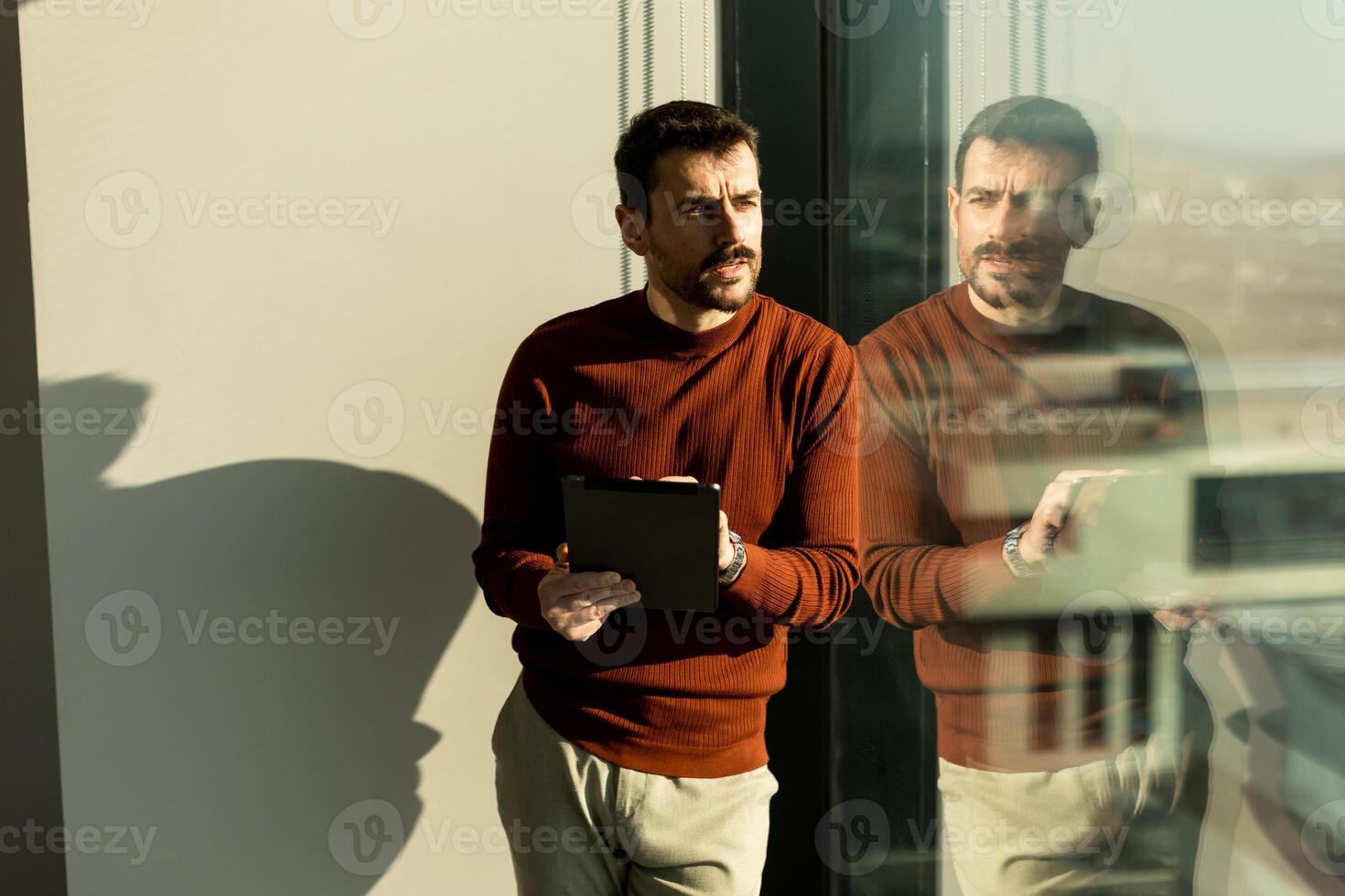 Professional man engages in deep thought while using a tablet next to a large window with city view photo