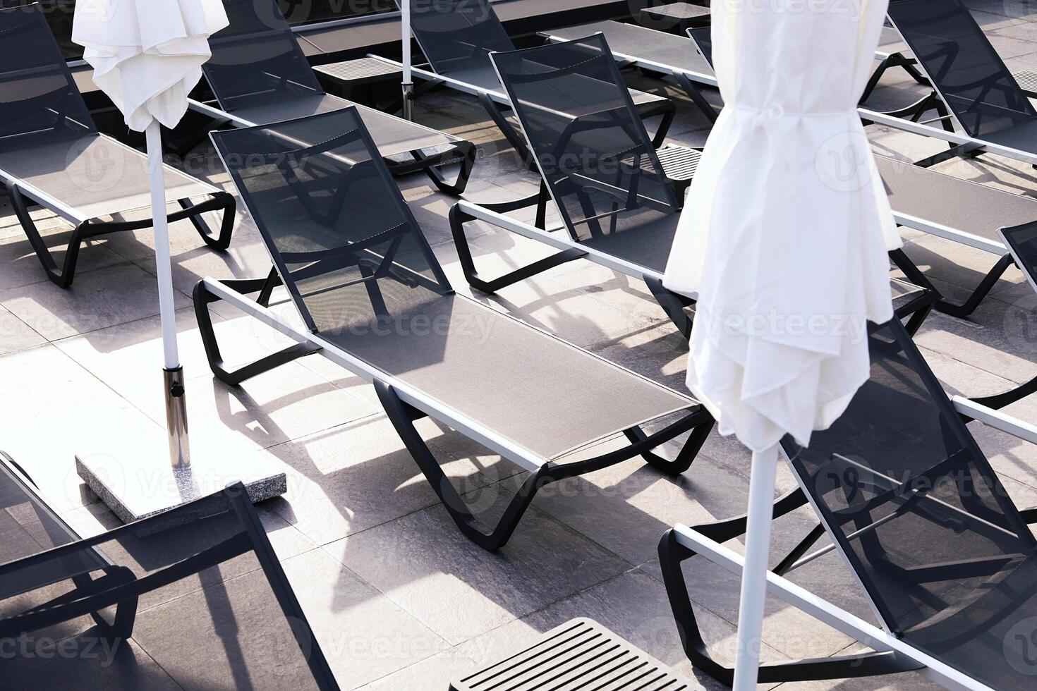 Modern sun loungers and closed umbrellas on a resort terrace, arranged in geometric patterns with sunlight and shadows photo