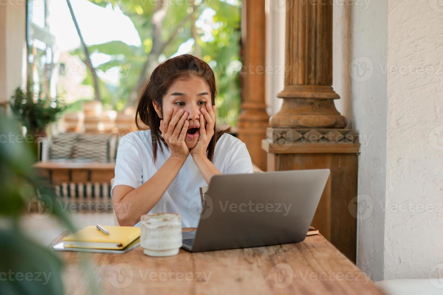 A woman looks surprised while working on a laptop in an outdoor setting with tropical plants and wooden columns. photo