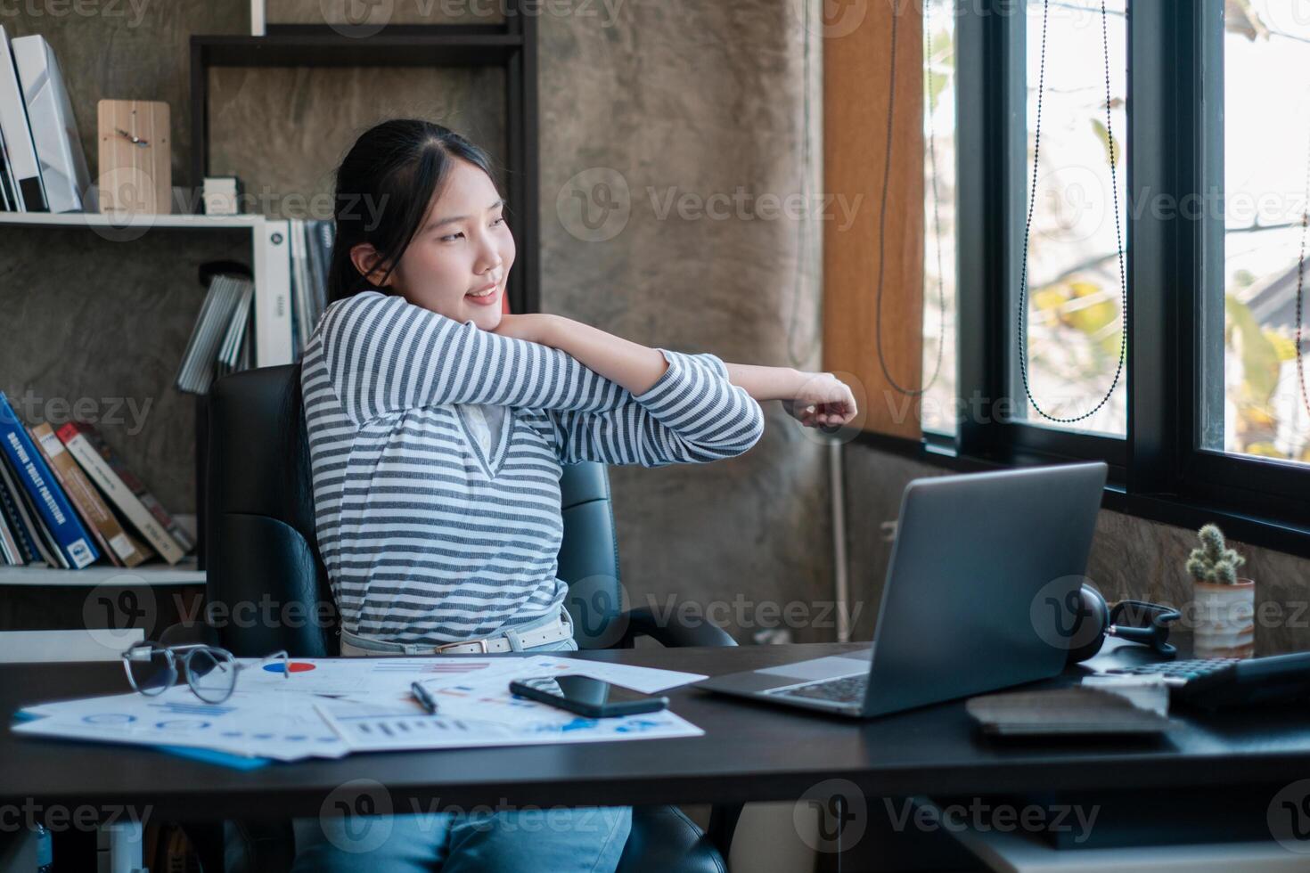 A young woman takes a break to stretch at her desk in a modern office setting, surrounded by work materials and a laptop. photo