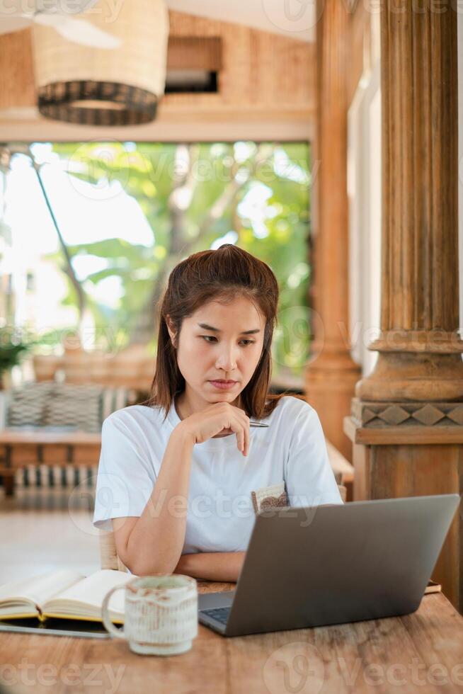 Focused woman using a laptop in a tropical outdoor space, surrounded by greenery, with a coffee mug and open notebook. photo