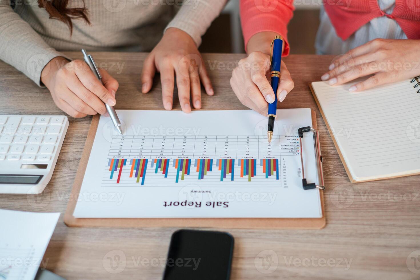 Two colleagues reviewing a sales report with colorful graphs, using pens and calculators on a wooden desk. photo