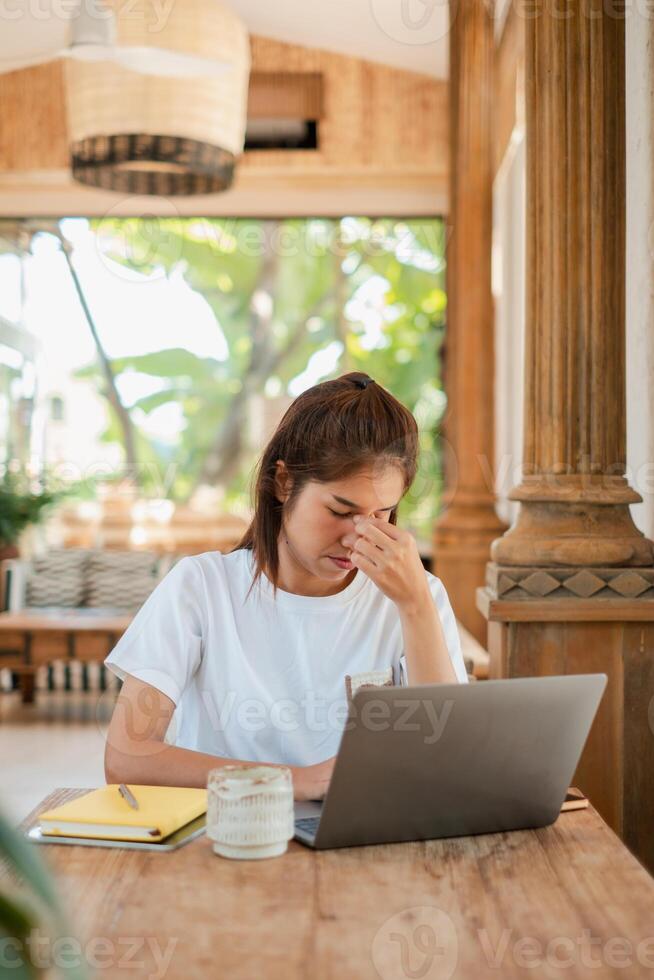 A woman in a white shirt looks stressed while working on a laptop in a tropical outdoor setting with wooden columns. photo