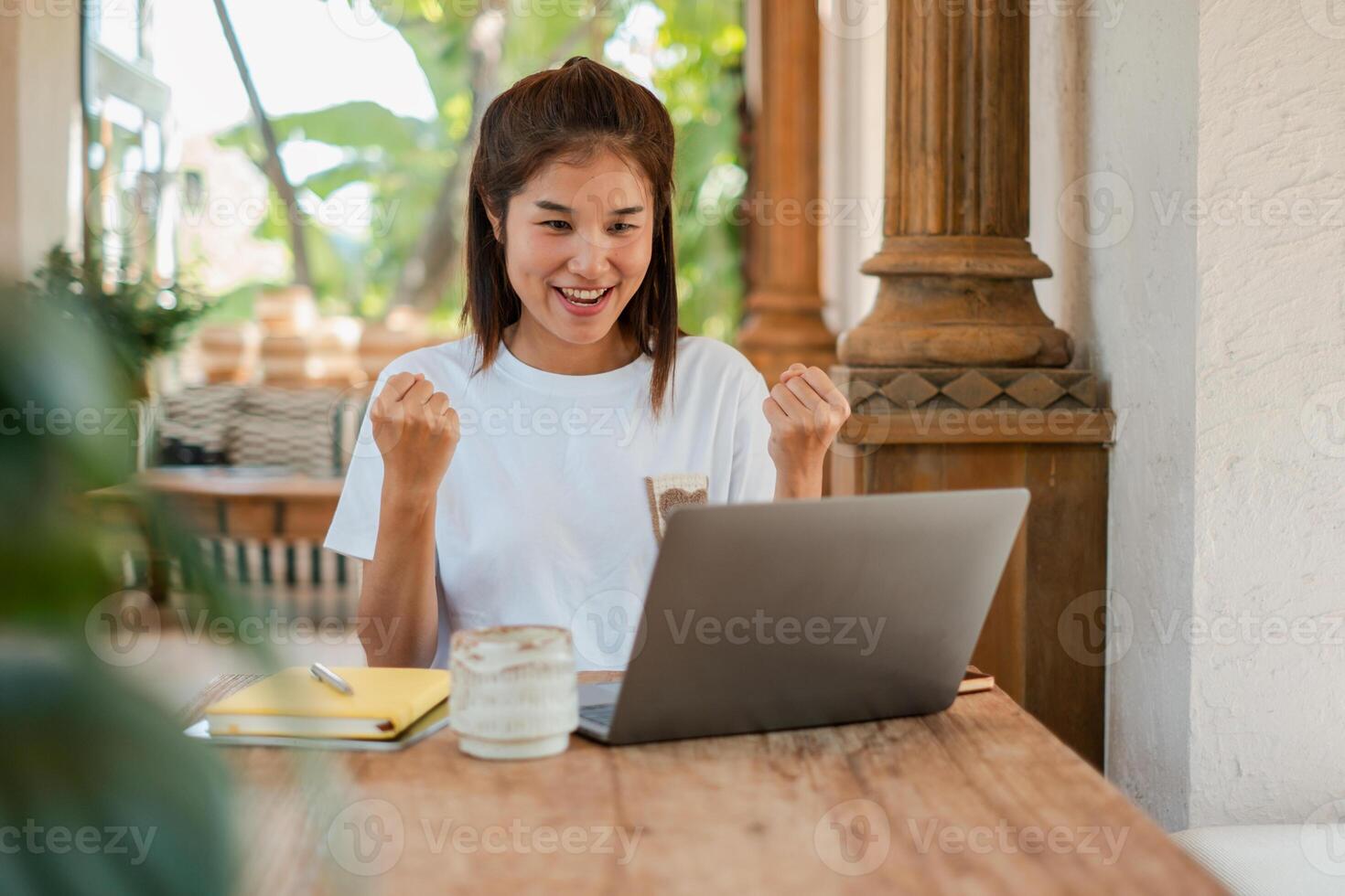 Joyful woman in casual attire celebrating success while using a laptop in a cozy outdoor setting with natural light. photo