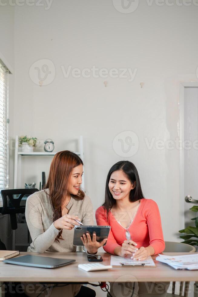 Two women discussing work in a bright office, using a tablet and surrounded by documents, showcasing teamwork and technology. photo
