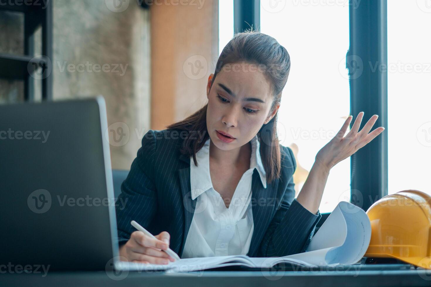 Professional woman in a suit working on documents at a desk with a laptop and construction helmet, showcasing focus and determination. photo