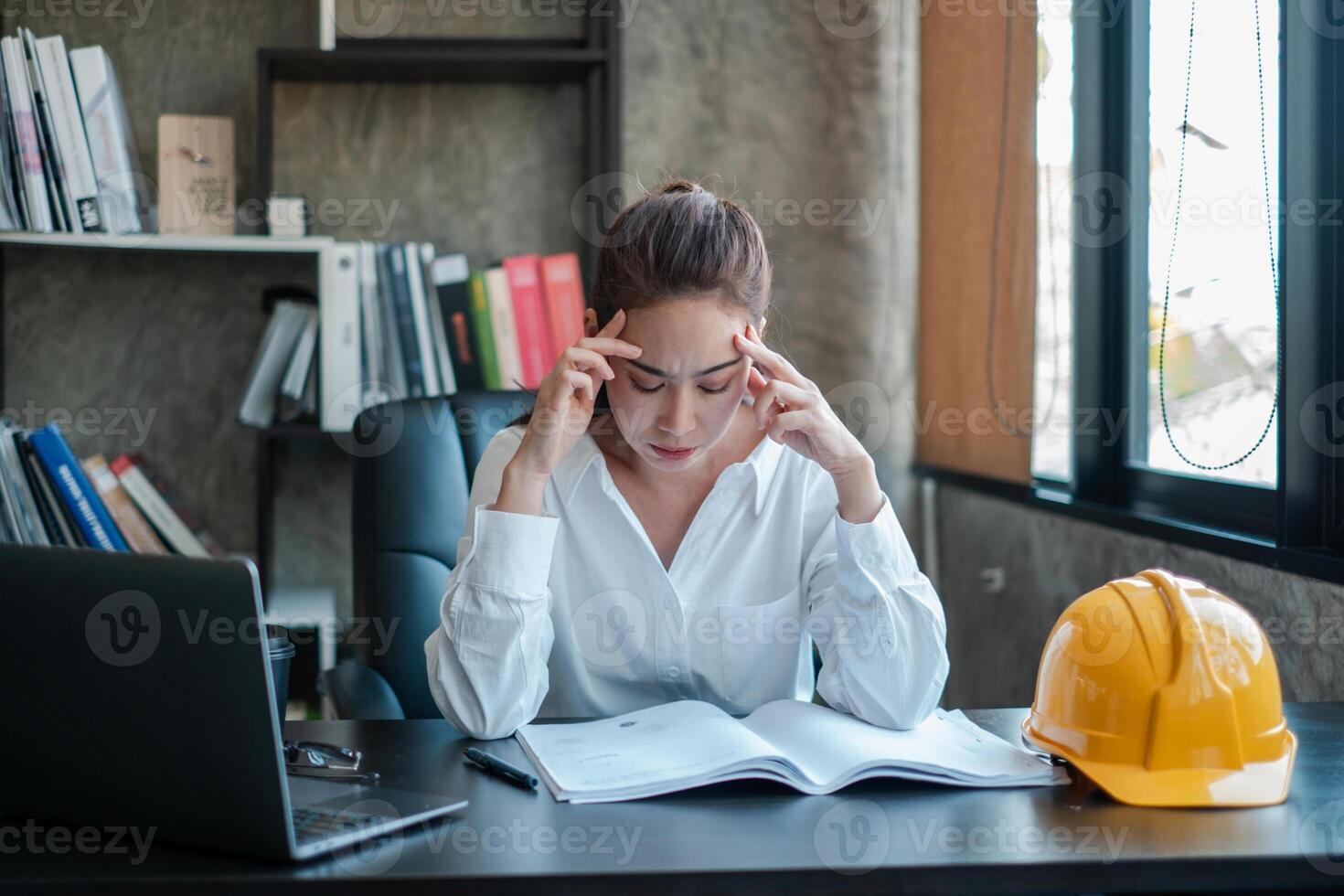 A female engineer appears stressed while working at her desk, surrounded by books and a safety helmet in a modern office environment. photo