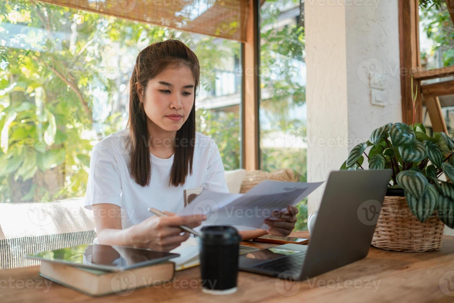 A young woman focuses on her work, surrounded by greenery and natural light, using a laptop and reviewing documents in a cozy home setting. photo