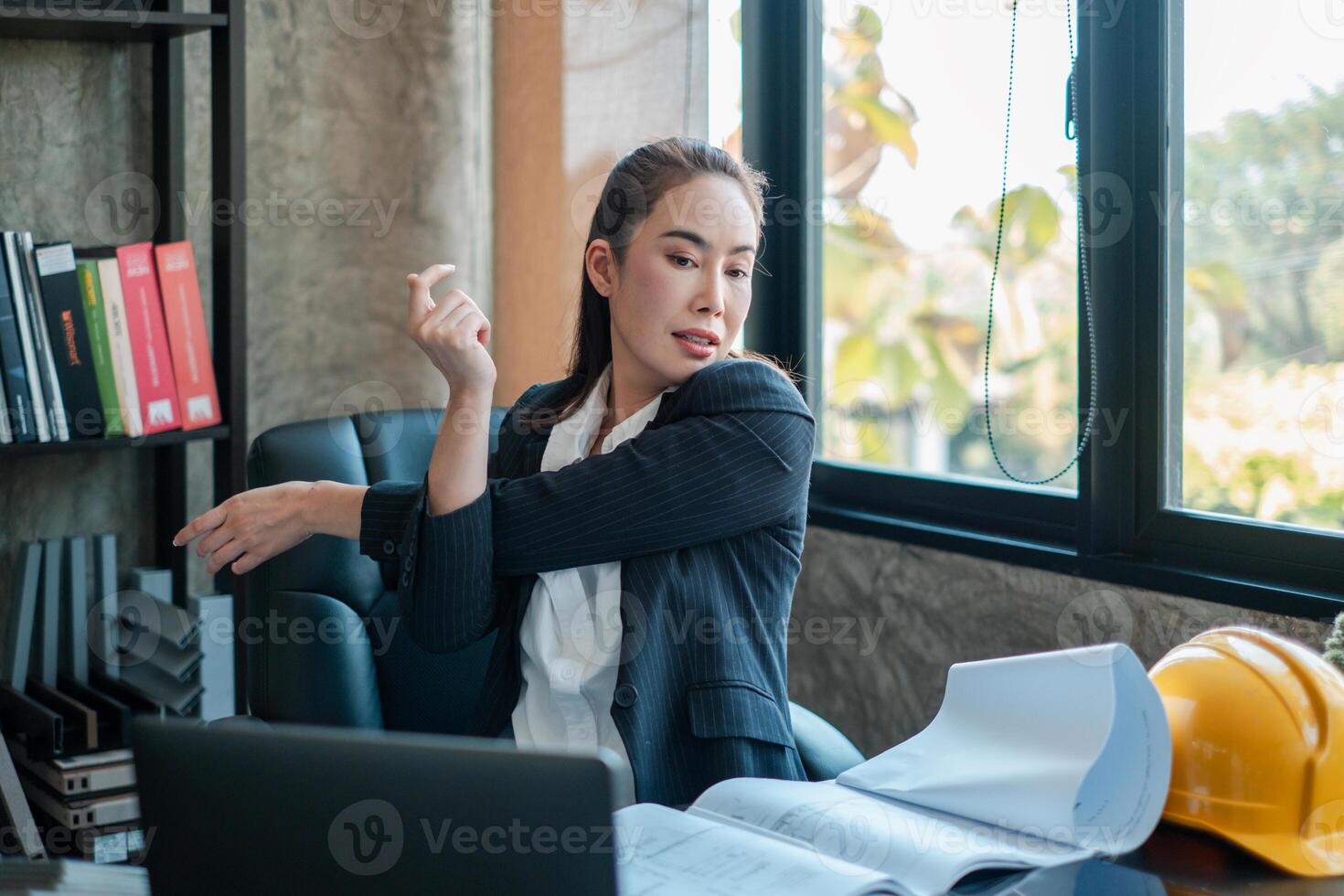 Professional woman in office attire stretching at her desk, surrounded by work materials and natural light from a large window. photo