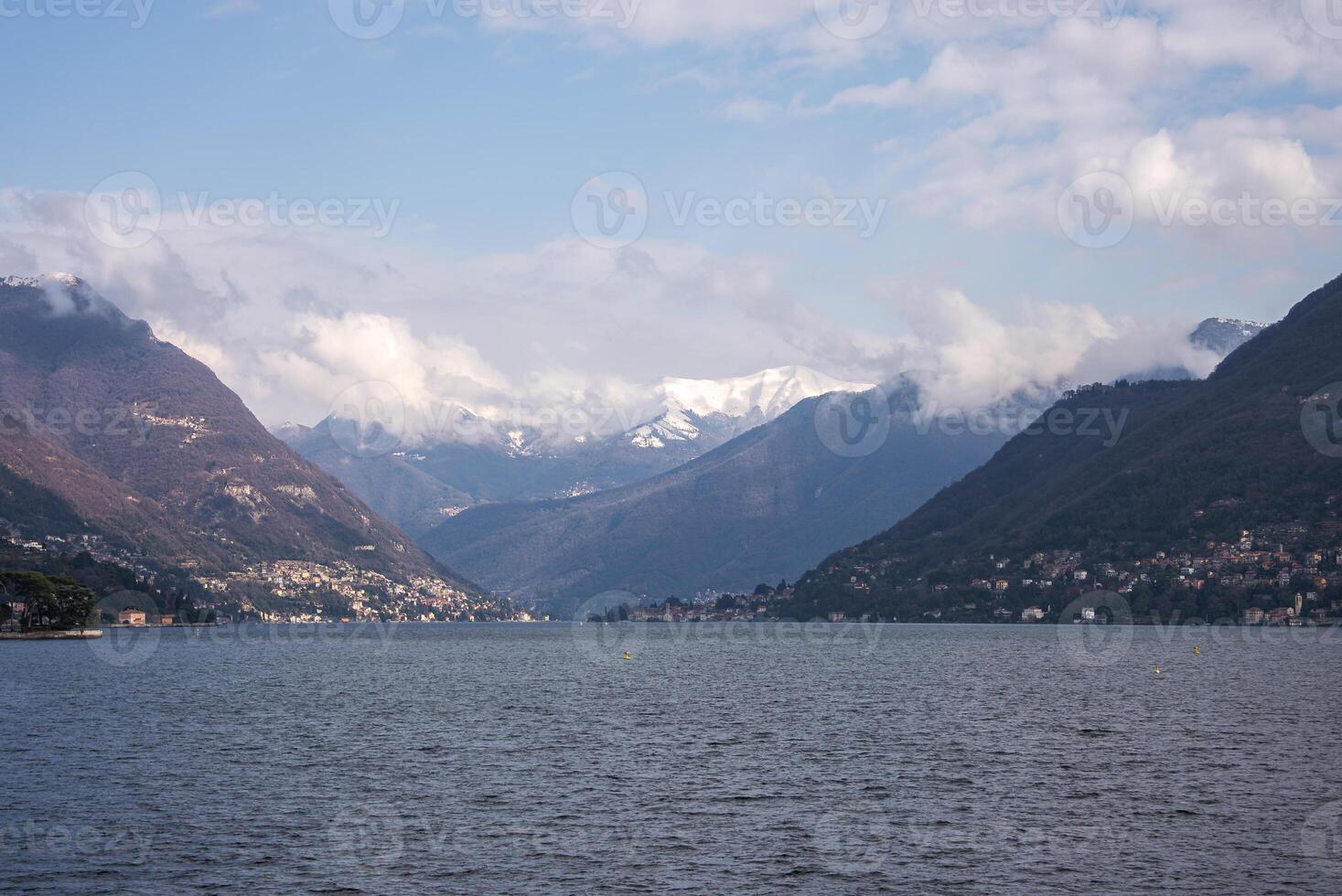 Lake Como is surrounded by forested hills and distant snow capped mountains. Small towns line the shoreline under a partly cloudy sky. photo