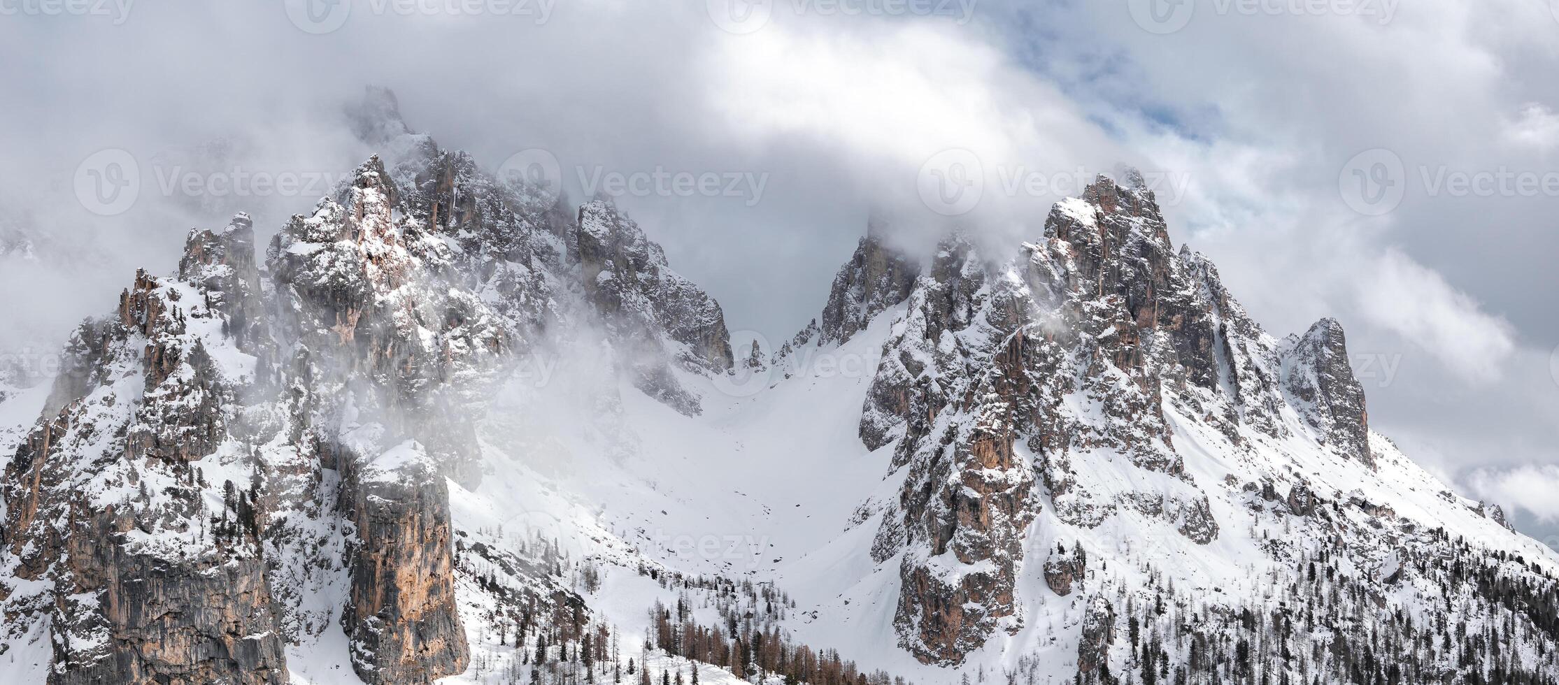 Snow covered Dolomite peaks near Cortina, Italy, partially shrouded in mist. Sparse trees dot the lower slopes, contrasting with the rugged terrain. photo