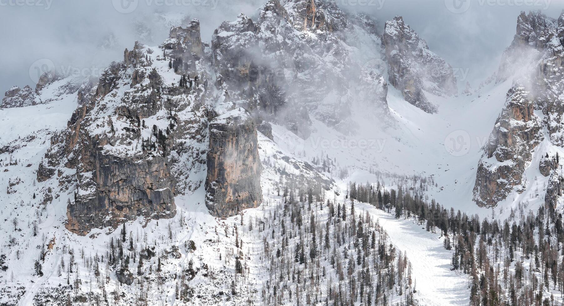 Snow covered peaks of the Dolomites in Cortina, Italy, with mist, rocky cliffs, and a ski slope lined with sparse, snow dusted trees in the foreground. photo