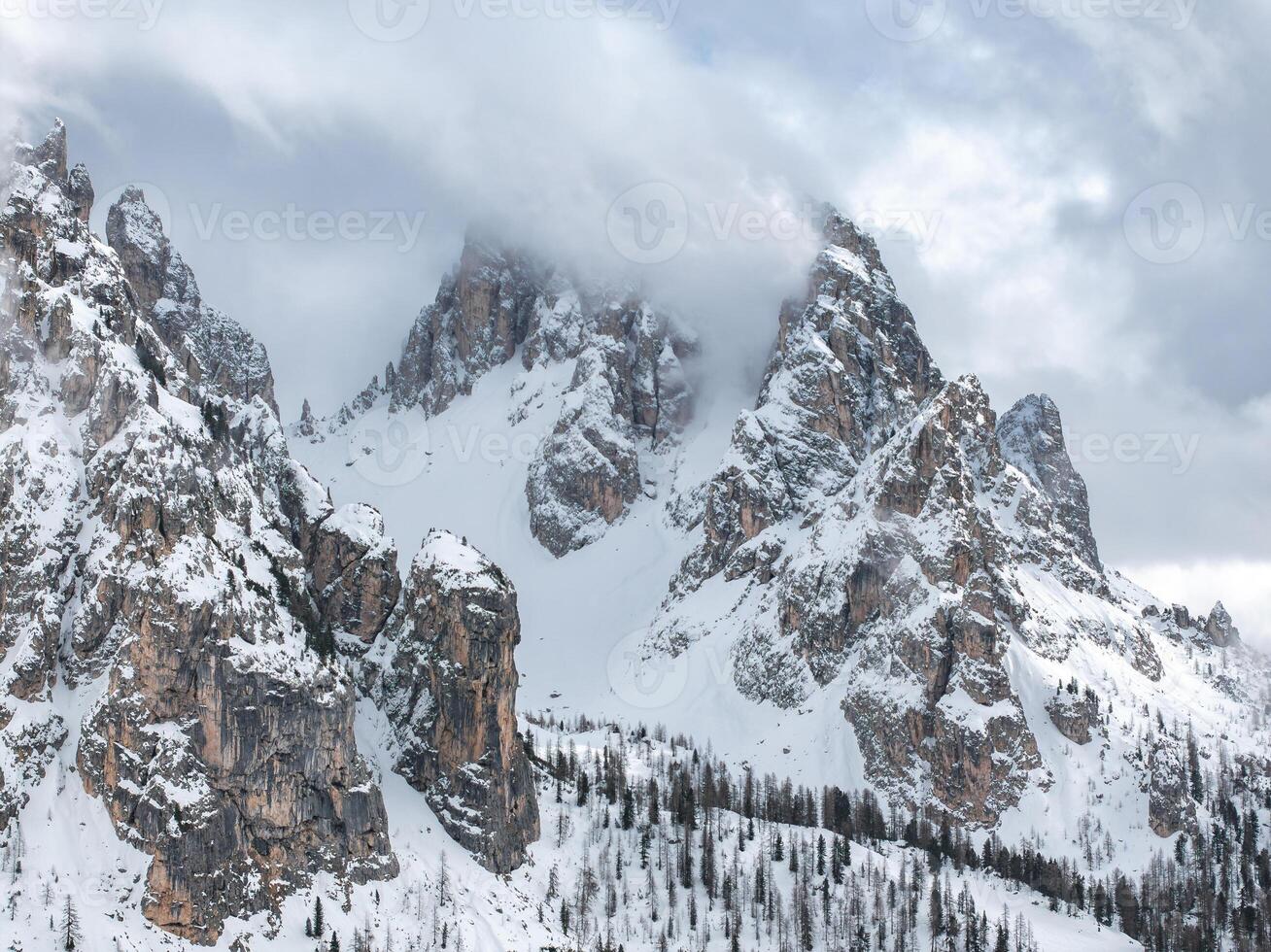 Snow covered Dolomite peaks near Cortina, Italy, partially shrouded in clouds. Sparse coniferous trees dot the lower slopes, contrasting the rugged terrain. photo