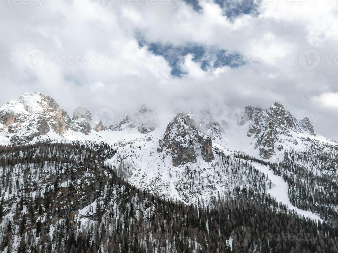 Snow covered peaks of the Dolomite Mountains in Italy rise above dense evergreen forests, with clouds partially shrouding the rugged terrain. photo