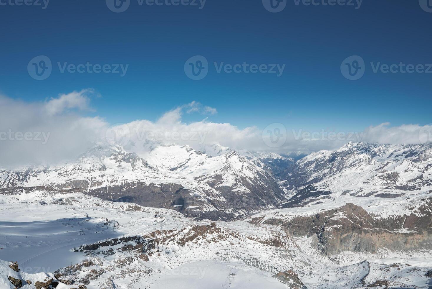 Snow covered alpine peaks and glaciers near Zermatt, Switzerland, under a clear blue sky with light clouds. Rugged terrain and valleys add dramatic contrast. photo