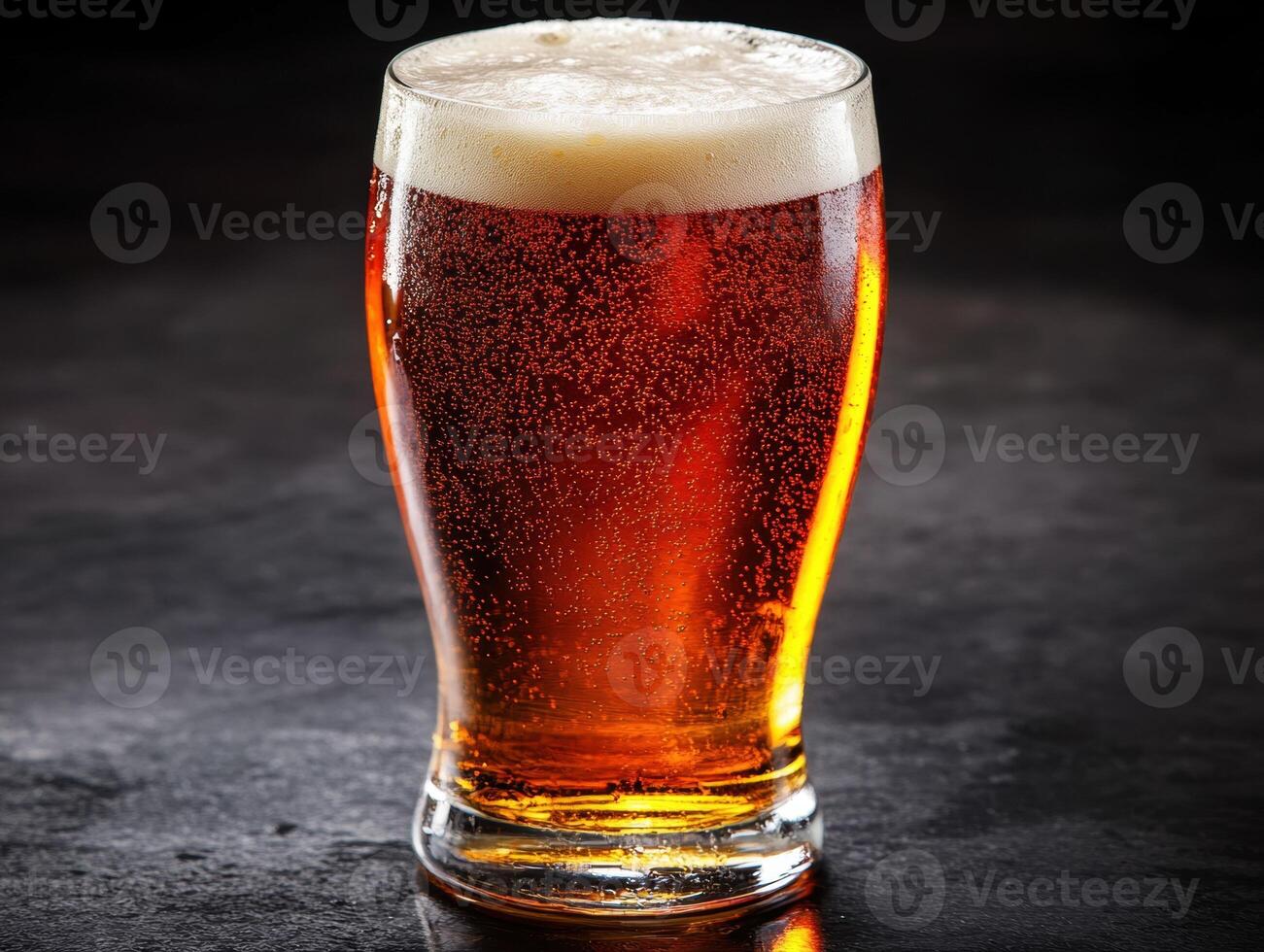 Sparkling amber beer in a clear glass container on dark surface photo