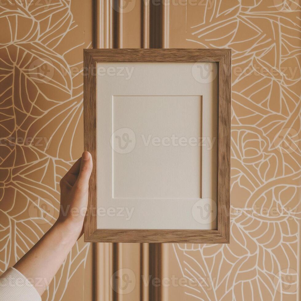 A person holds up an empty, wooden picture frame against a patterned brown and white background photo