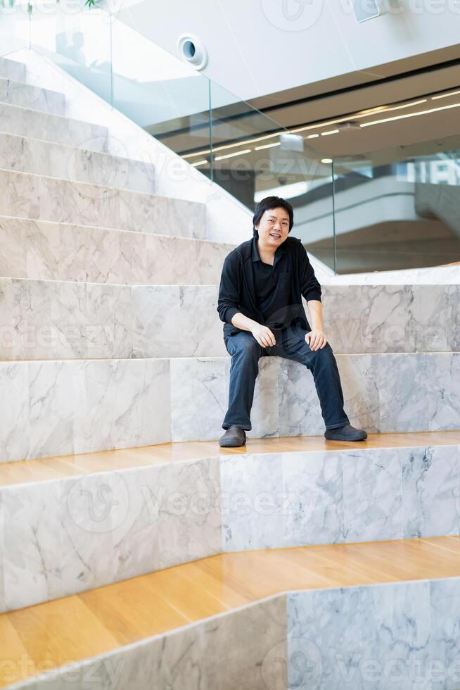 Asian Man in Black cardigan and polo shirt sits on the 3white grand marble stairs in the grandstand with wood floor on its step. photo