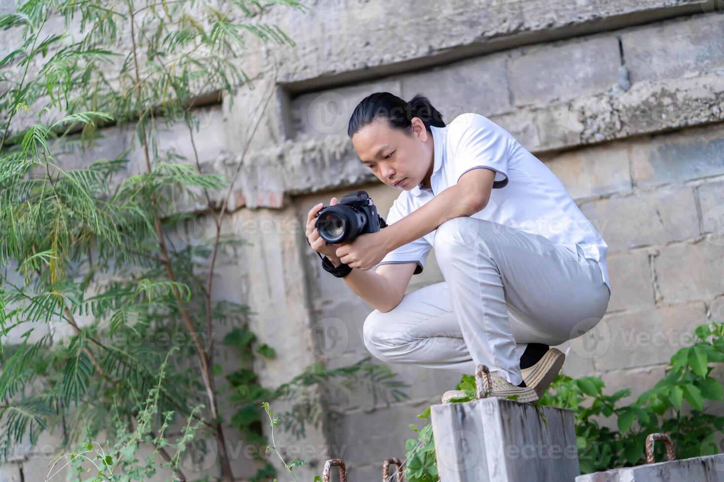 Professional Asian Chinese - Thai Camera man posture 9and focus in viewfinder and LED screen on mirrorless camera medium format in construction concrete area. photo