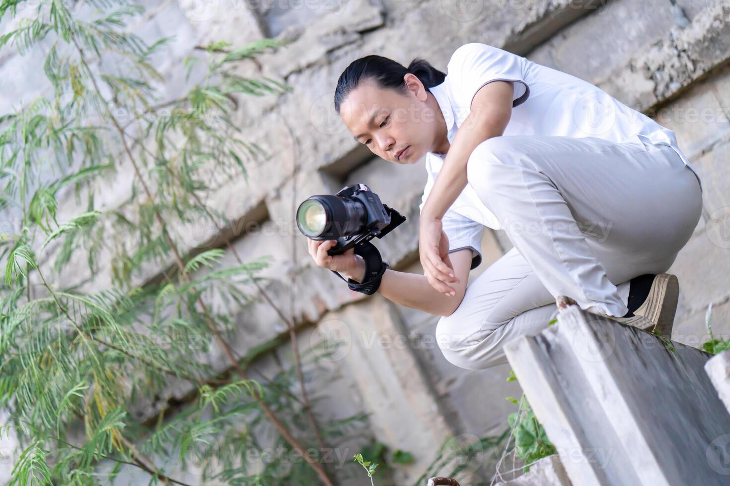 Professional Asian Chinese - Thai Camera man posture 6and focus in viewfinder and LED screen on mirrorless camera medium format in construction concrete area. photo