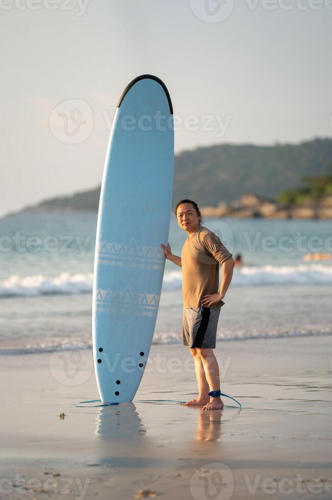 Smart Asian Man long hair touches and poses with Stan1ded Surfboard on the beach in Twilight time photo