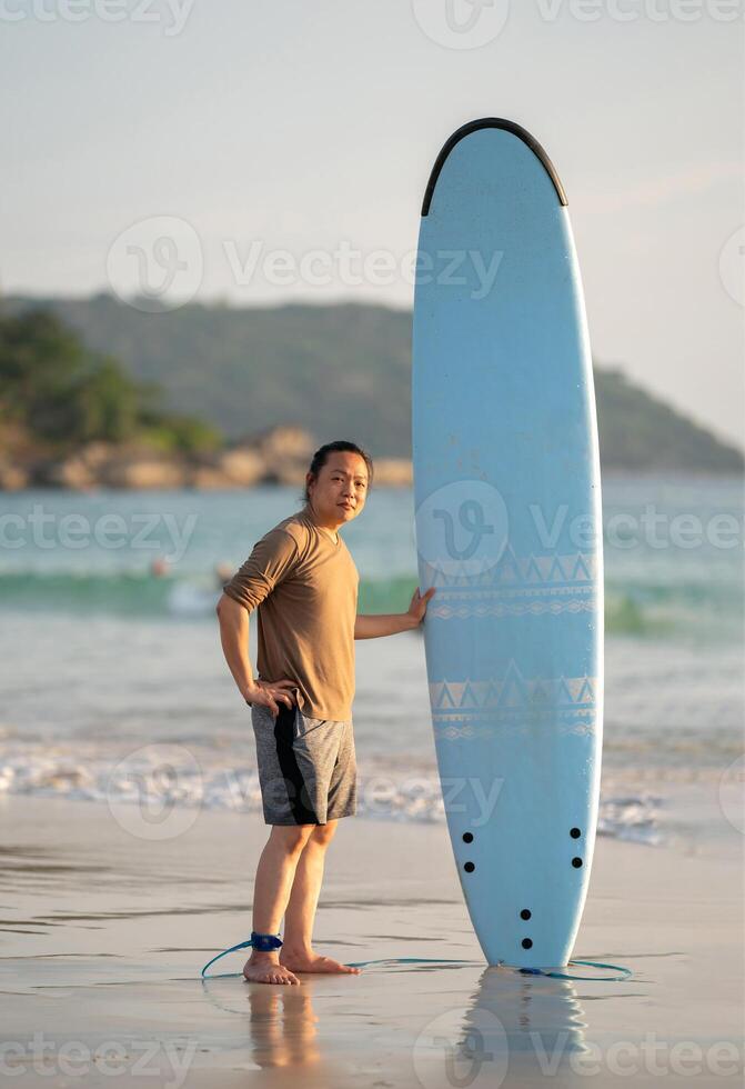 Smart Asian Man long hair touches and poses with Standed Surfboard on the beach in Twilight time1 photo