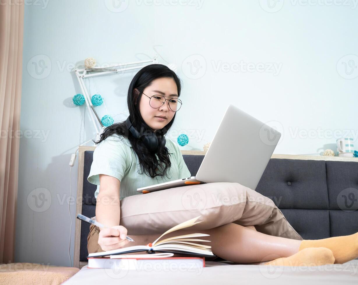 Glasses beautiful Asian woman is working from home with her laptop and write a note into notebook beside in the bedroom with a pastel green - brown color theme.55 photo