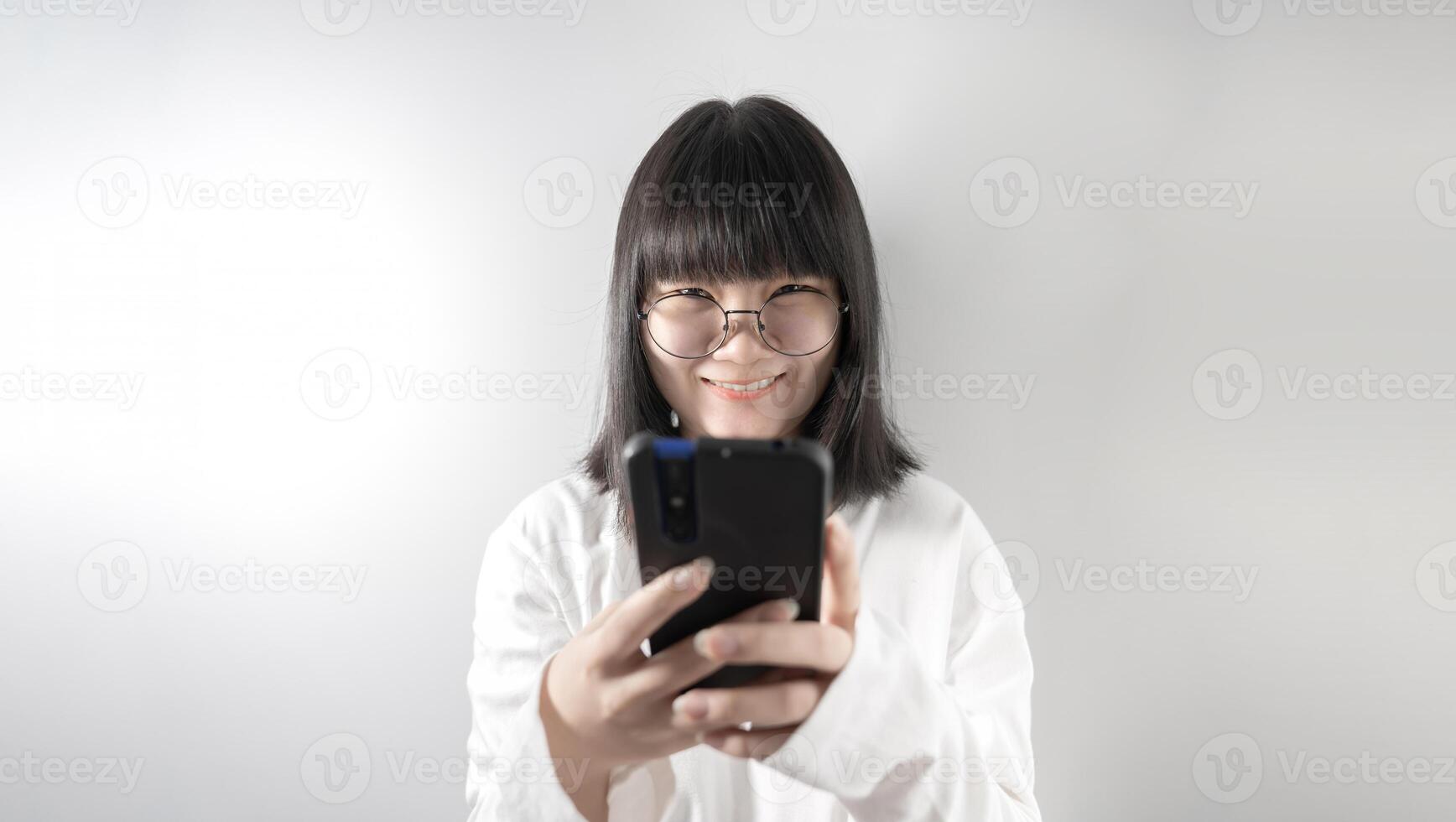 Isolated Pretty Asian glasses woman looks at camera a1nd holds mobile phone in her hand on white background in studio light. photo