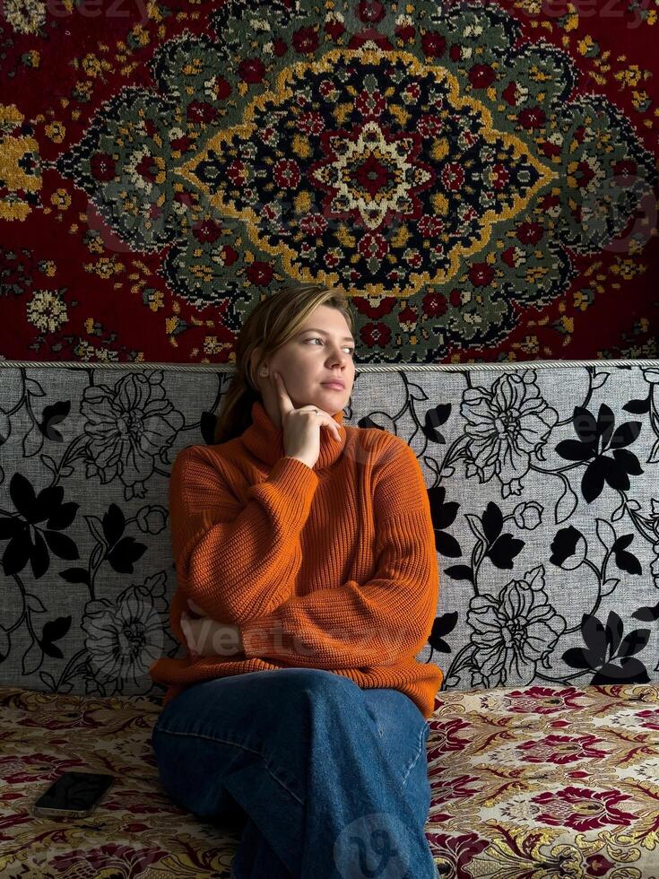 In a traditional Russian home, a woman wearing a cozy orange sweater sits on a patterned couch, with a richly decorated vintage carpet displayed on the wall behind her. photo