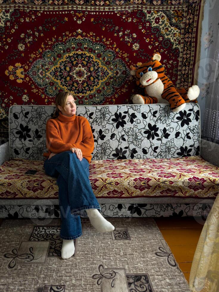 A woman in a cozy orange sweater sits on a patterned couch in a traditional Russian home, with a richly decorated vintage carpet hanging behind her. photo
