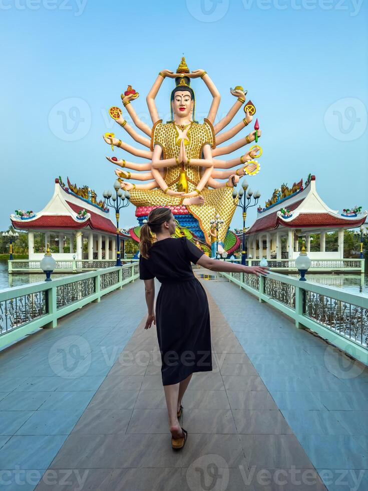 A woman walks along a bridge towards the grand, multi-armed statue of the Goddess Guanyin in Thailand. The statue's vibrant colors contrast with the clear blue sky, creating a striking visual photo