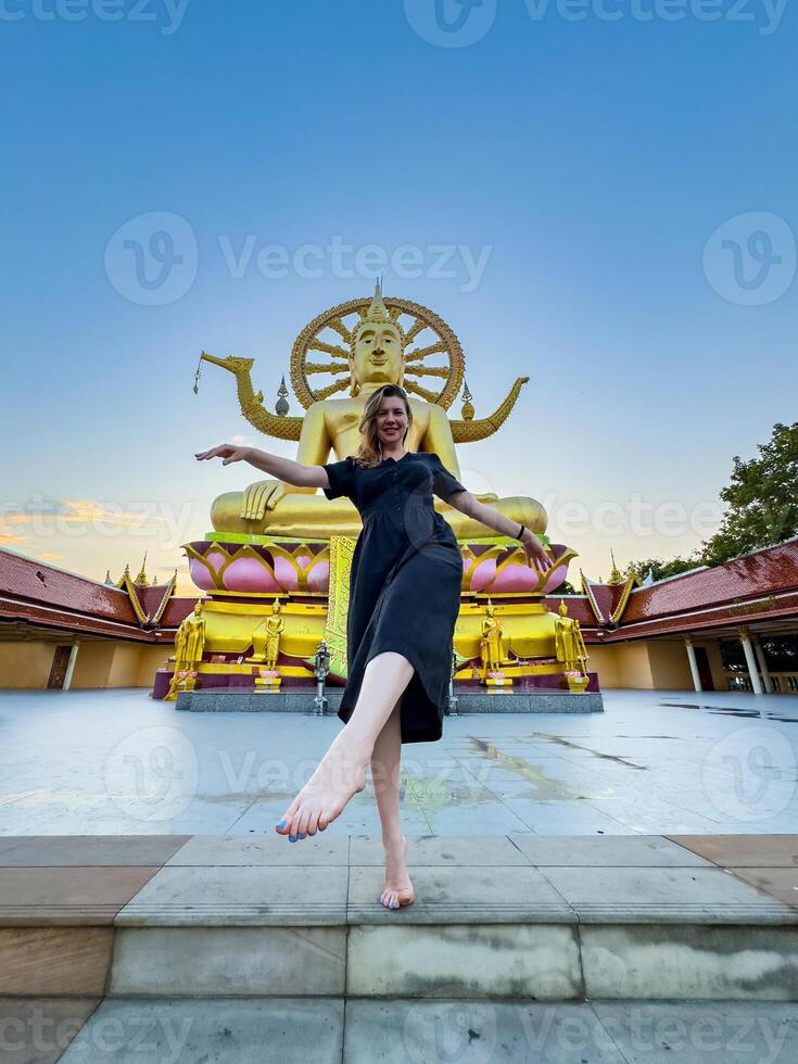 A young woman in a black dress joyfully poses barefoot on temple steps, aligning her arms with the golden Big Buddha statue at sunset. Shot from a low angle for a dramatic effect photo
