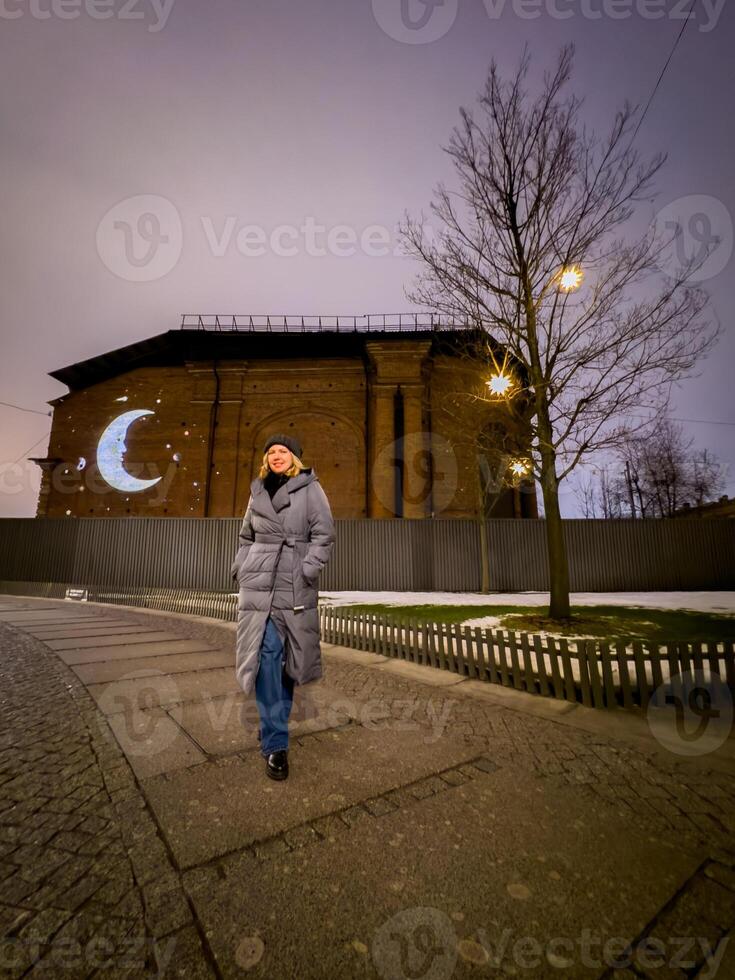On a cold winter evening, a woman in a long gray puffer coat walks through the city. A glowing crescent moon and stars are projected onto an old building wall, adding a dreamy photo