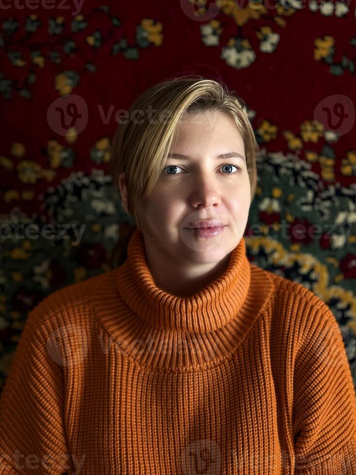 A woman clad in a comfortable orange sweater is seated on a patterned sofa in a traditional Russian dwelling, a lavishly ornamented vintage rug adorning the wall behind her. photo