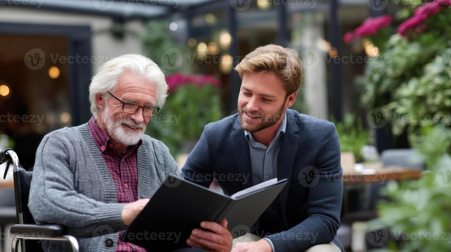Elderly man in wheelchair sharing photo album with younger man, both smiling in serene garden setting