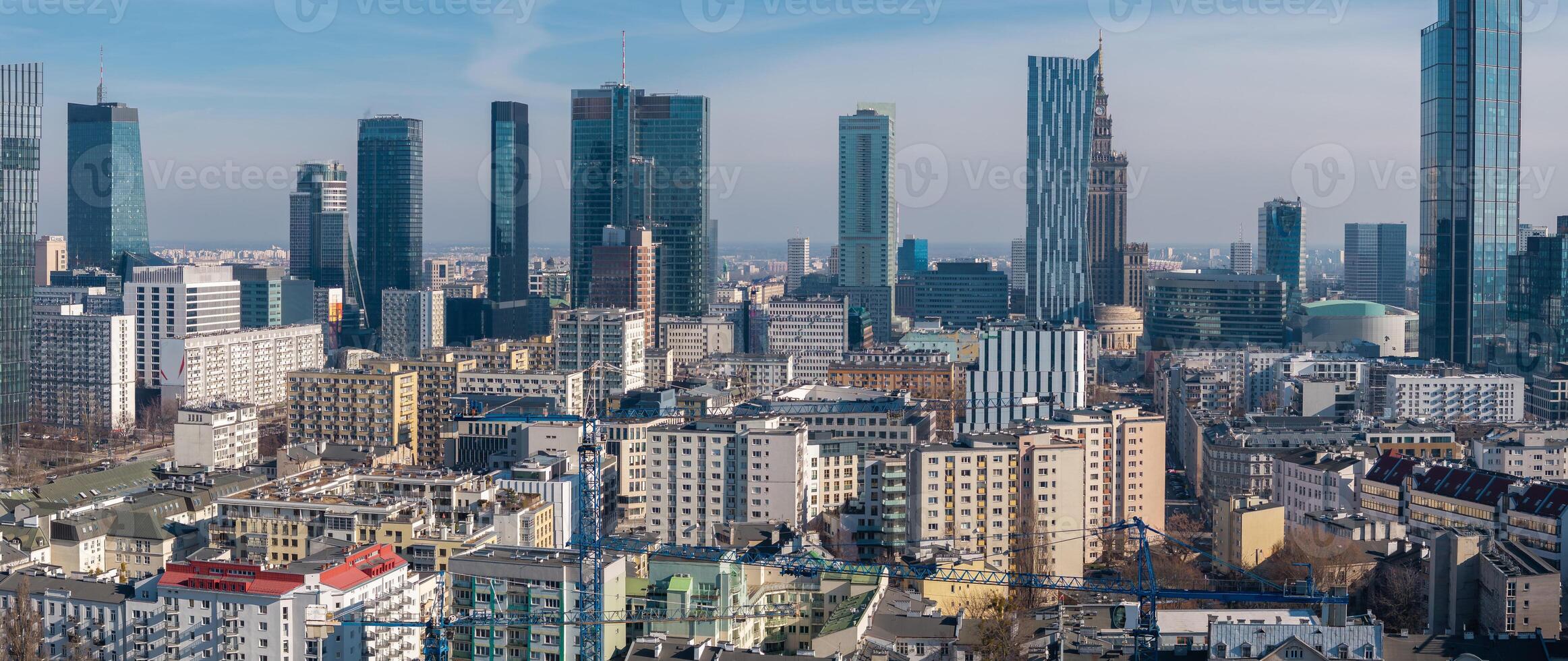 Aerial view of Warsaw, Poland, showing skyscrapers, the Palace of Culture and Science, cranes, and a mix of modern and mid century buildings under a clear sky. photo