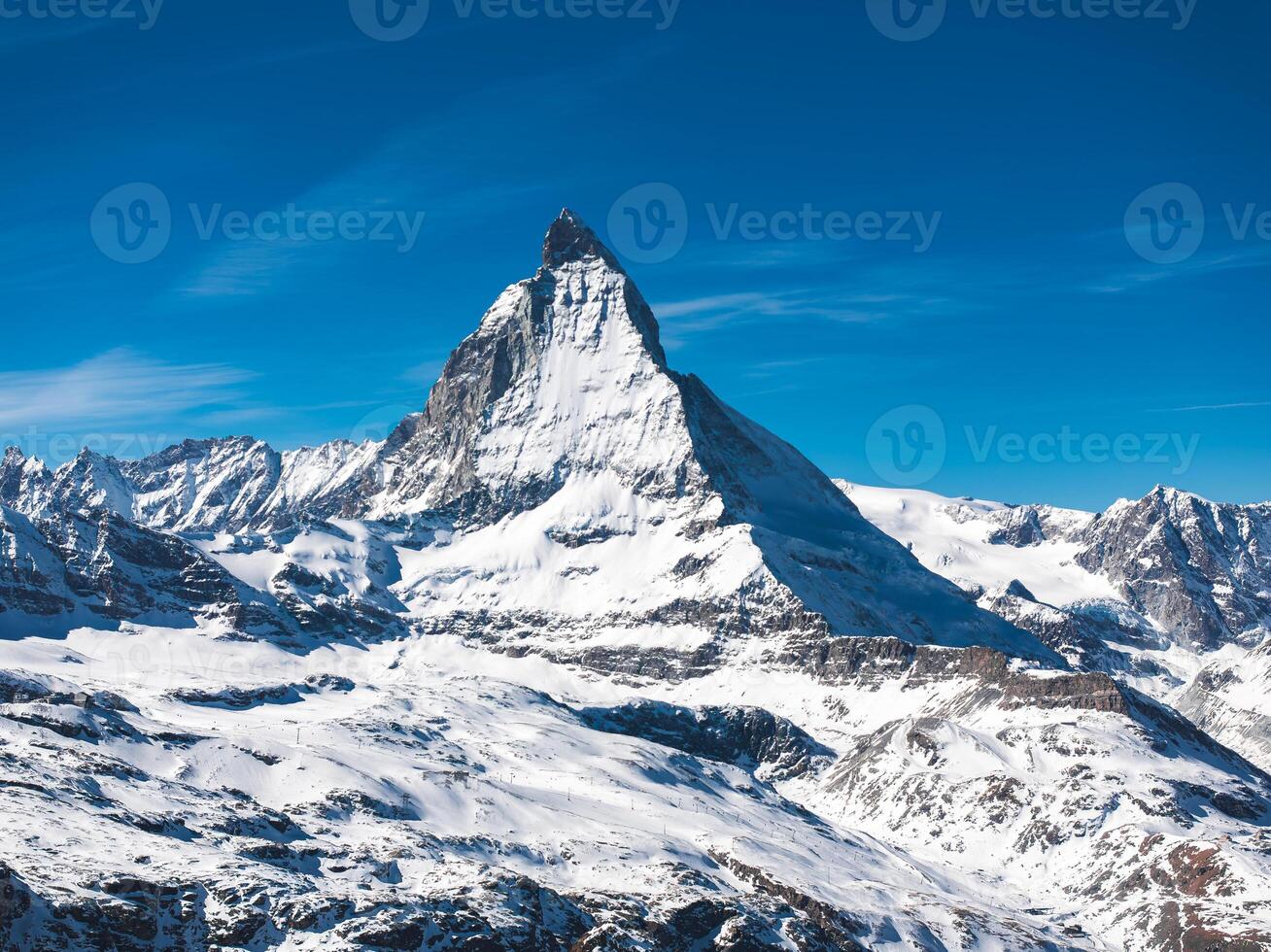 Aerial view of the iconic Matterhorn mountain in the Pennine Alps, surrounded by snow covered slopes and rugged alpine terrain under a blue sky. photo