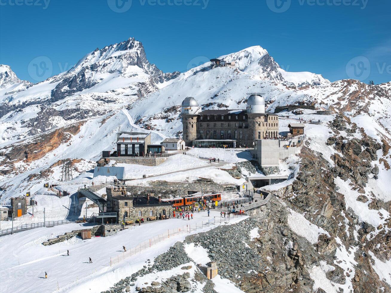 Aerial view of Gornergrat Observatory in Zermatt, Switzerland, with twin domes, snow covered Alps, Matterhorn peak, train station, and visitors below. photo