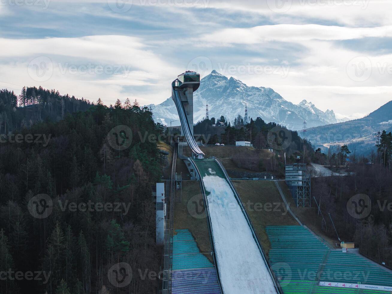 The Bergisel Ski Jump in Innsbruck, Austria, features a modern design, forested surroundings, and snow capped Alpine peaks in the background. photo
