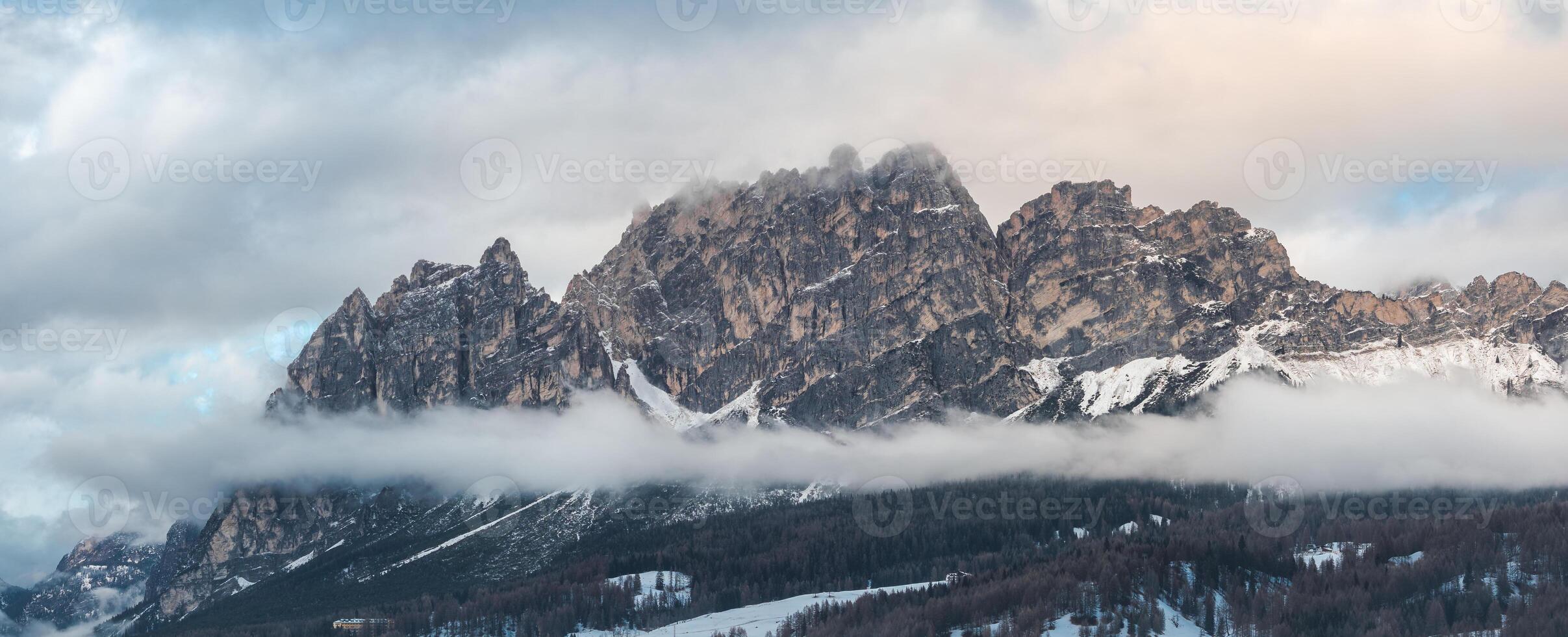 Aerial view of rugged Dolomite peaks near Cortina d'Ampezzo, Italy, with snow covered slopes, low clouds, and visible ski trails and structures. photo