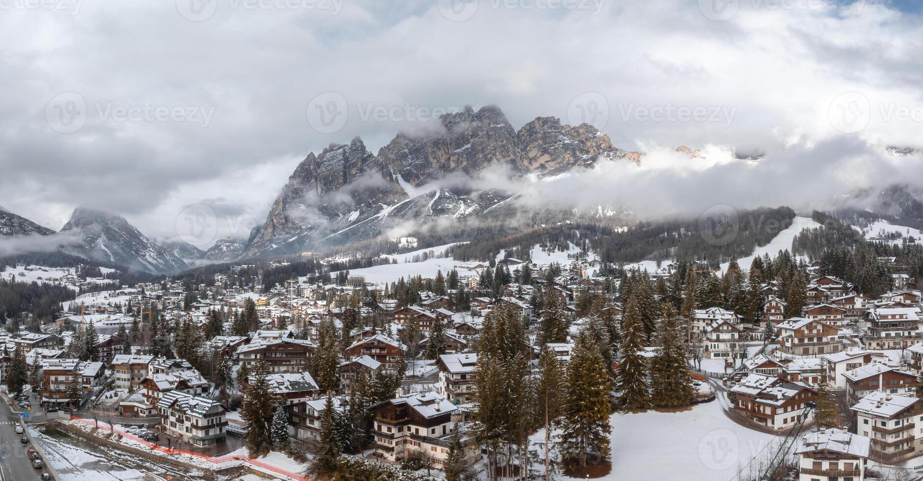 Aerial view of Cortina d'Ampezzo in Italy with snow covered rooftops, alpine buildings, evergreen trees, and Dolomites peaks partially in clouds. photo