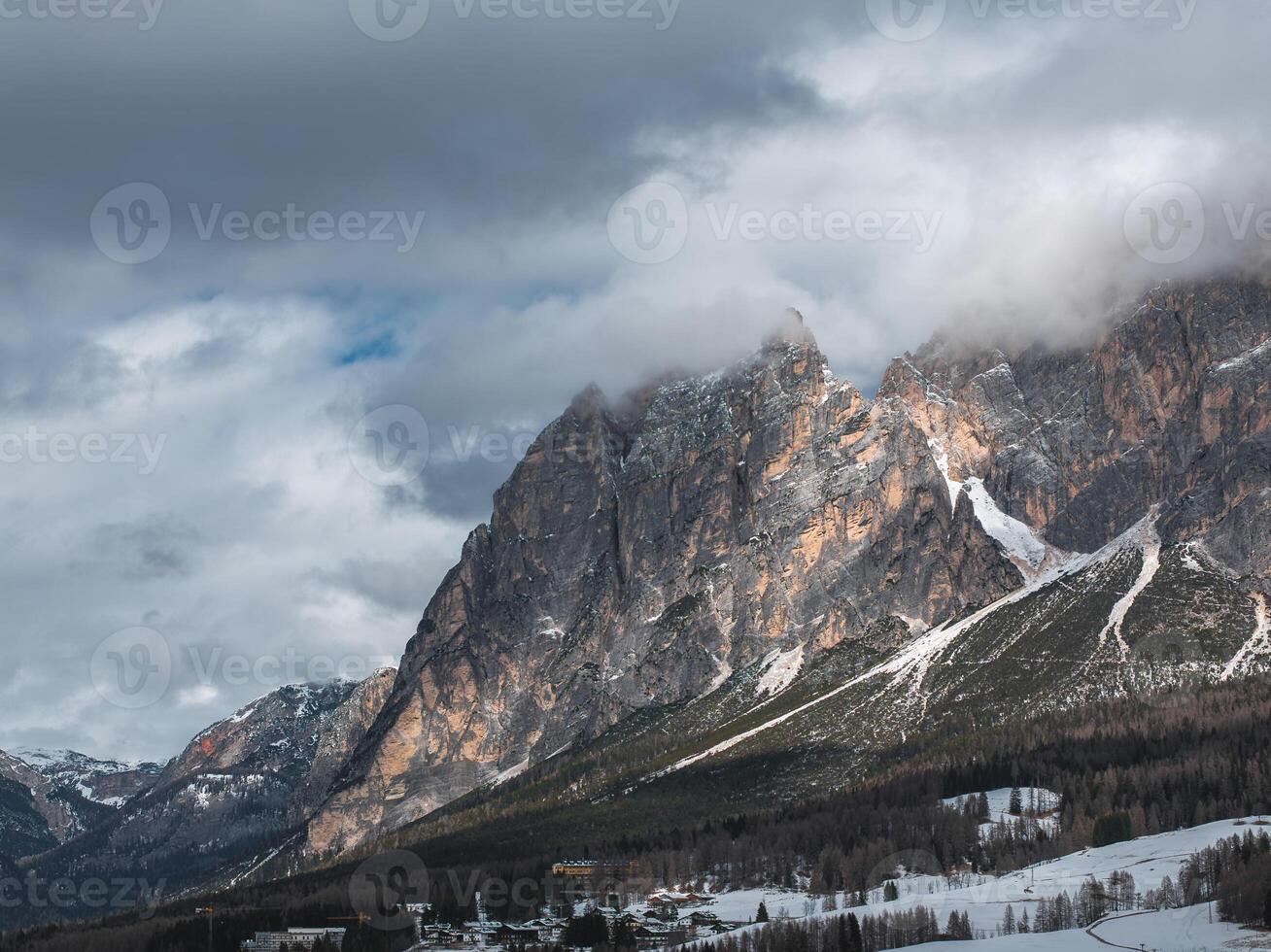 Cloud covered Dolomite peaks near Cortina d'Ampezzo, Italy, with sunlight on cliffs, a small town, forested areas, and snow patches below. photo