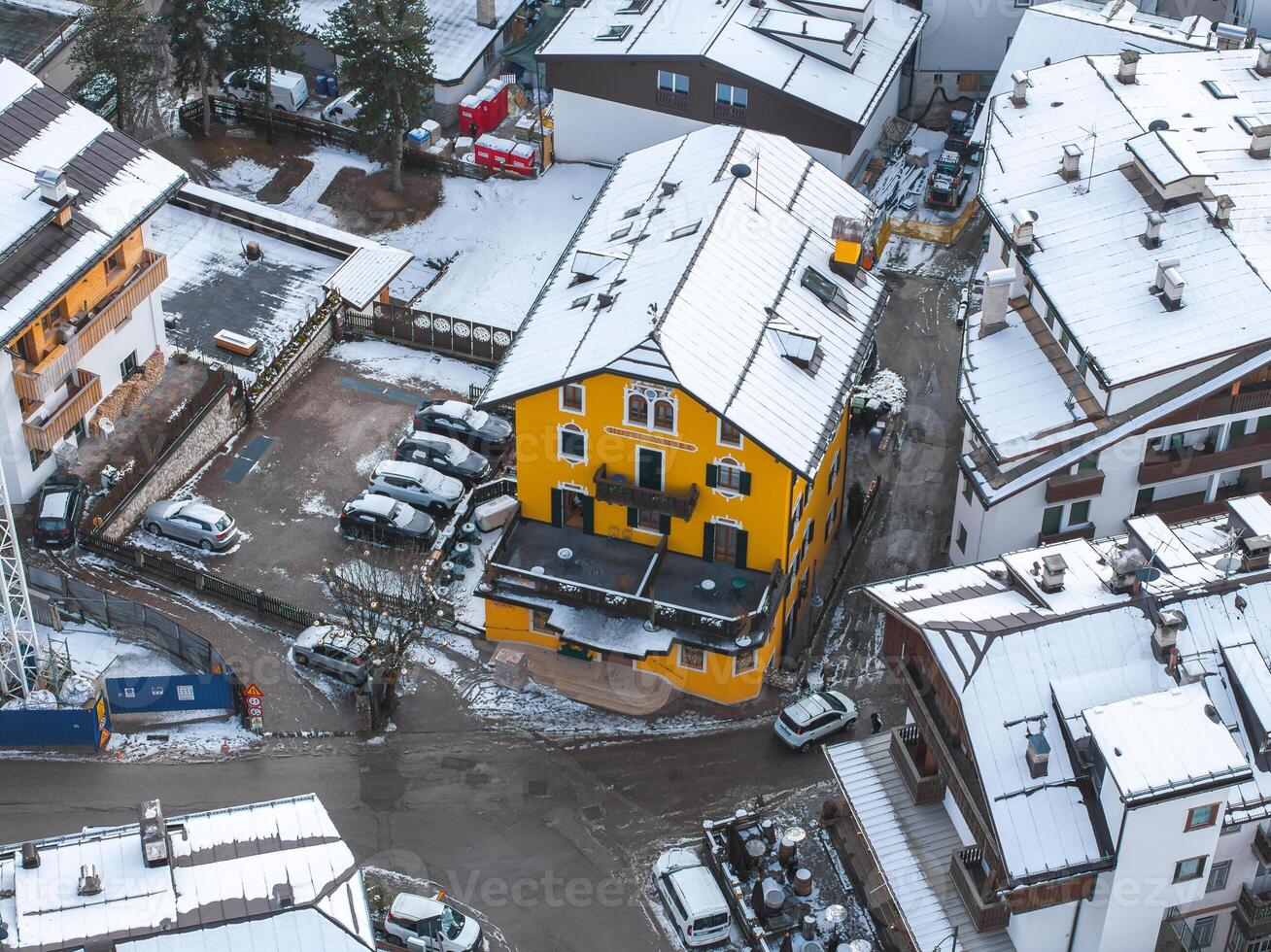 Aerial view of Cortina, Italy, featuring a bright yellow building with a sloped roof and arched windows, surrounded by alpine architecture and snowy rooftops. photo