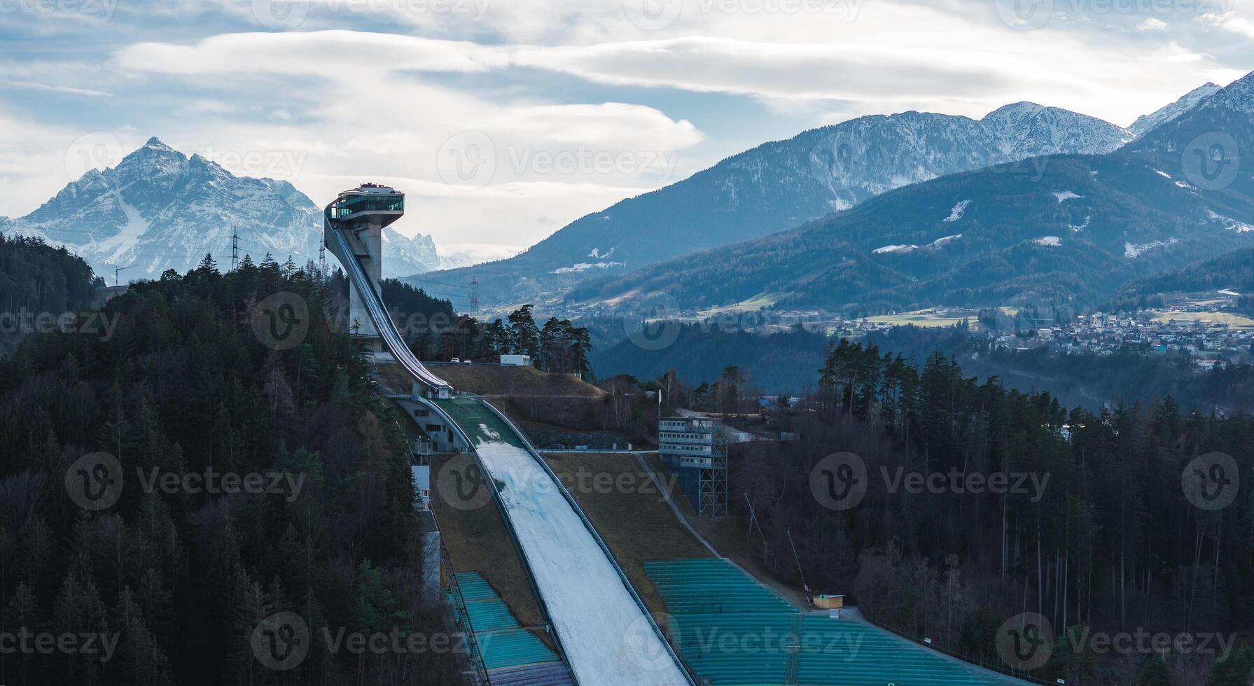 The Bergisel Ski Jump in Innsbruck, Austria, stands on a forested hill with its modern design, surrounded by dense forests and alpine mountain ranges. photo