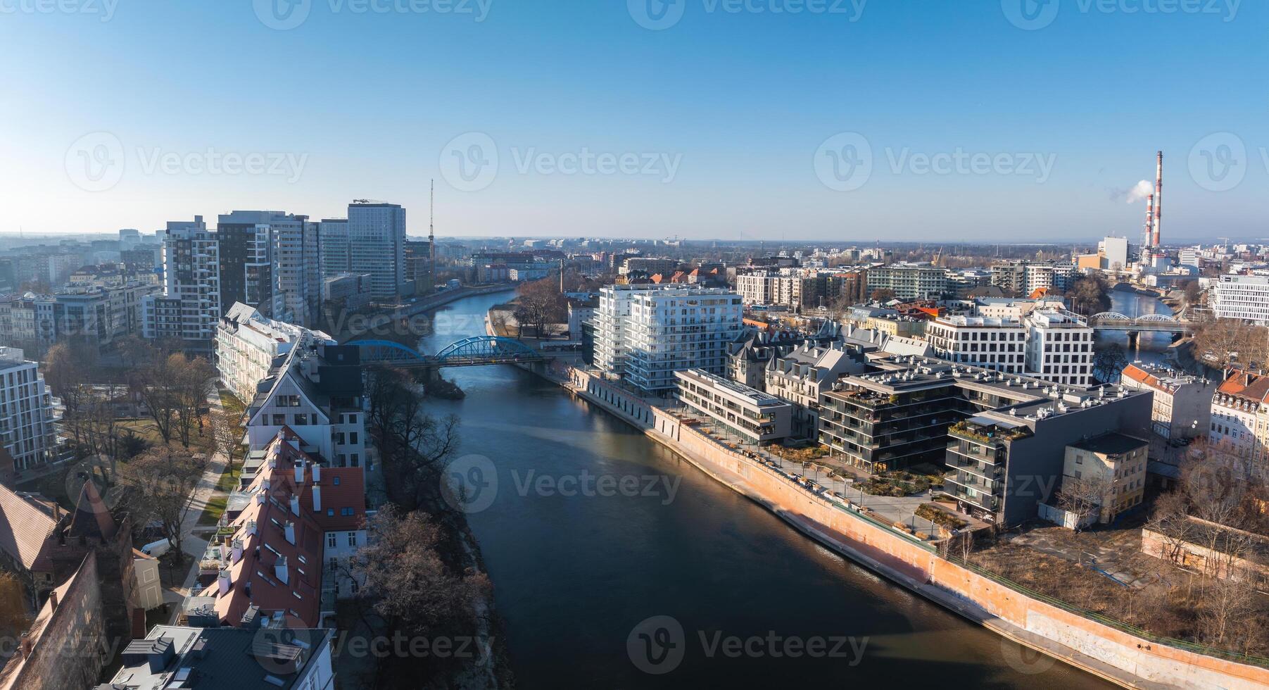Aerial view of Wroclaw, Poland, showing the Oder River, a prominent bridge, red roofed buildings, modern structures, and a tall industrial chimney. photo